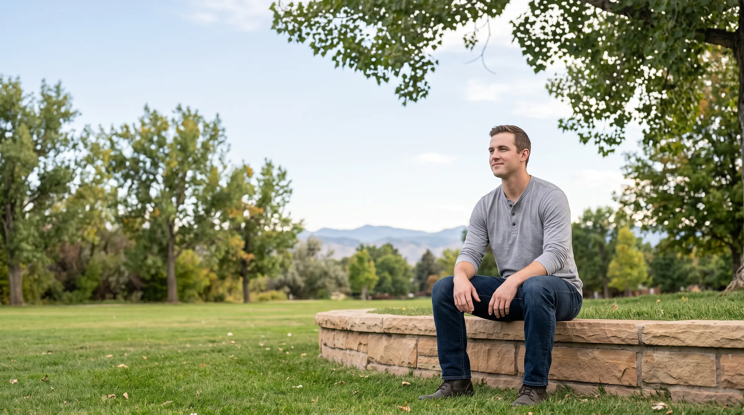 A man sitting on a low sandstone wall at the edge of a Front Range park, cottonwoods and foothills soft behind him.