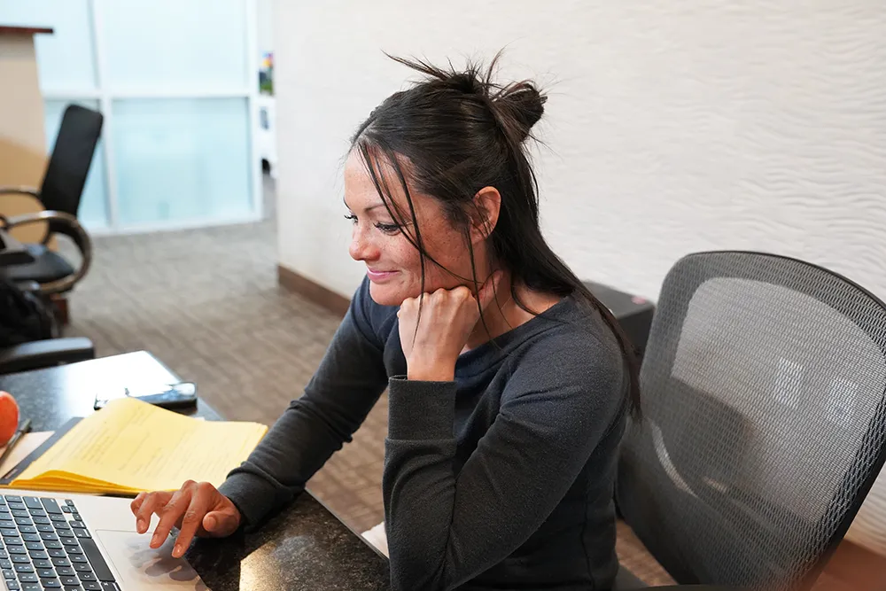 A staff member working at a desk with an open laptop, a yellow legal pad, and a mesh task chair.