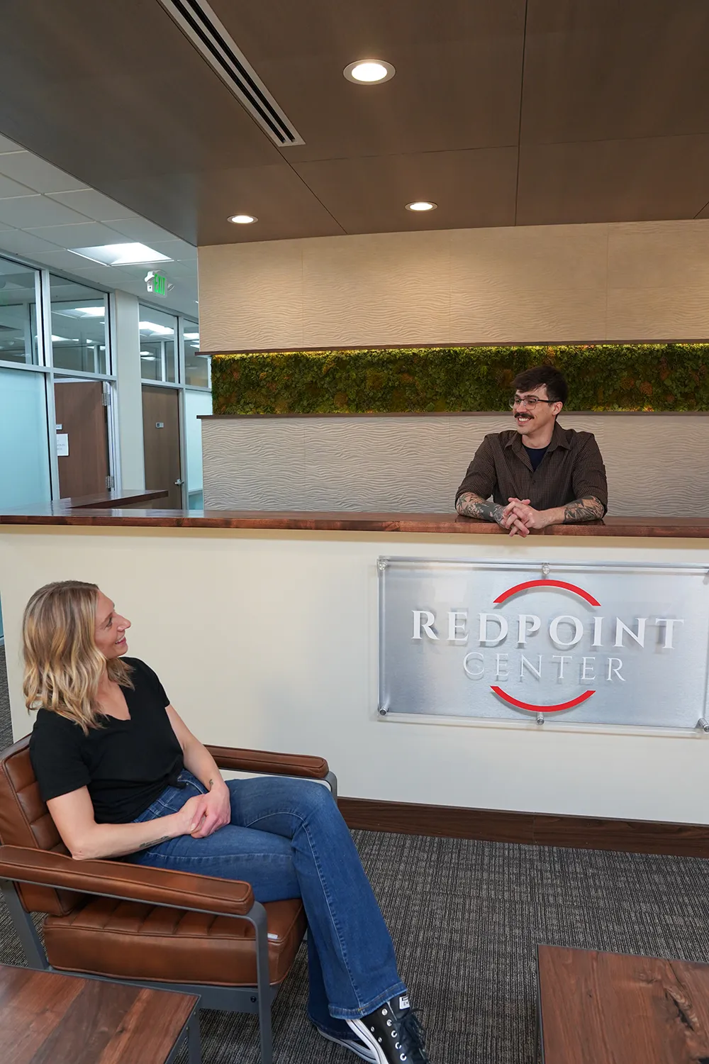 A woman seated in a tan leather chair in reception, turned toward a staff member leaning on the desk, with the moss wall and the REDPOINT CENTER sign behind them.