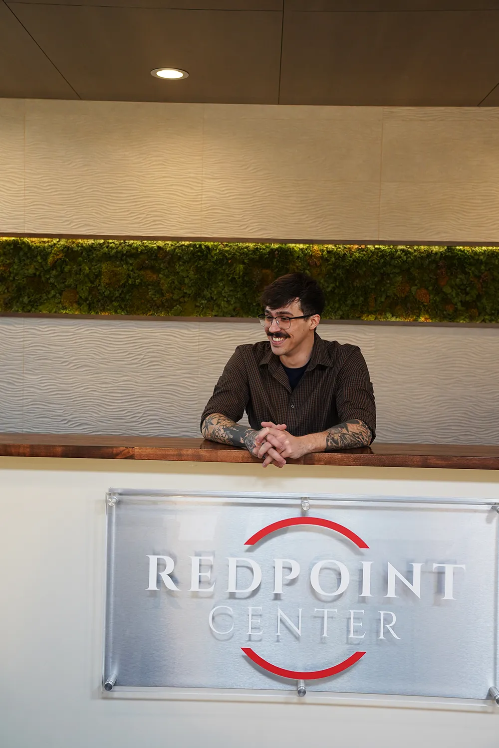 A staff member smiling behind the reception desk with hands clasped, above the brushed REDPOINT CENTER sign, the preserved moss wall behind.