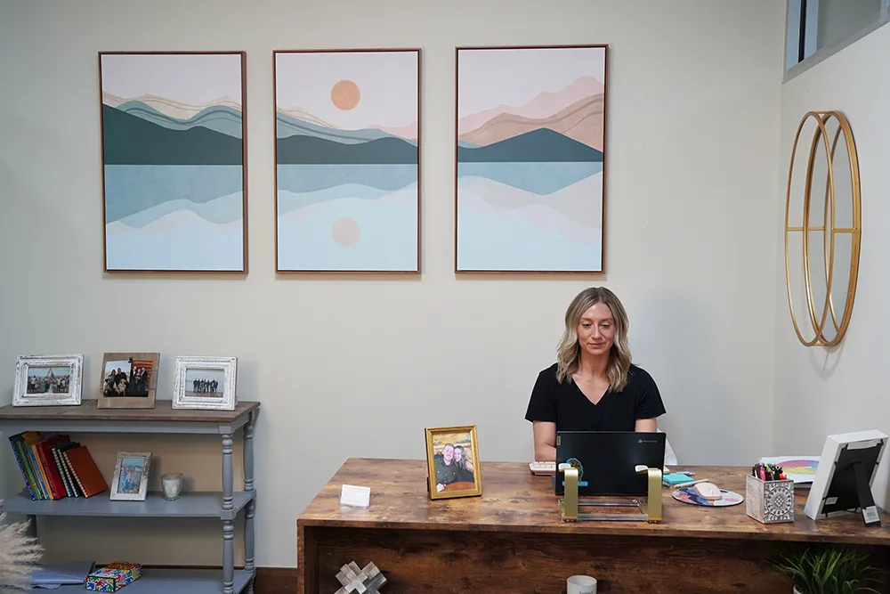 A front office with three framed mountain prints, open shelving of framed photos, and a staff member at a wood desk.