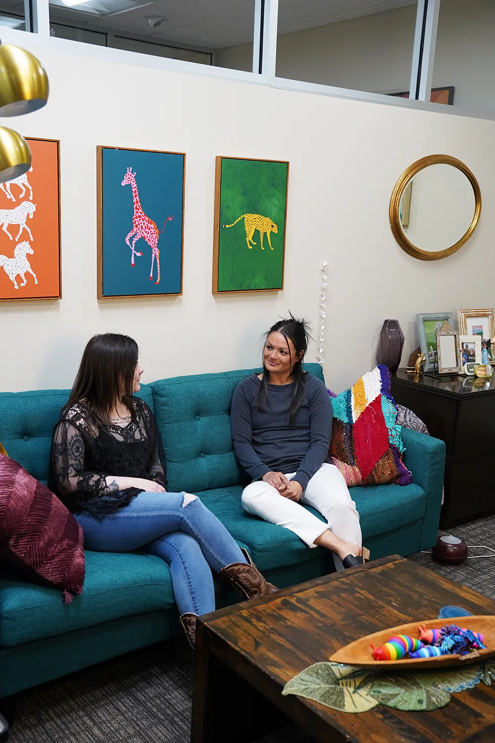 Two women in conversation on a teal tufted sofa beneath a triptych of painted animals, with gold pendant lights and a round gold mirror.