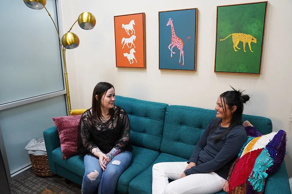 The same lounge head-on: a teal sofa under the animal triptych, a gold floor lamp, and two people talking.