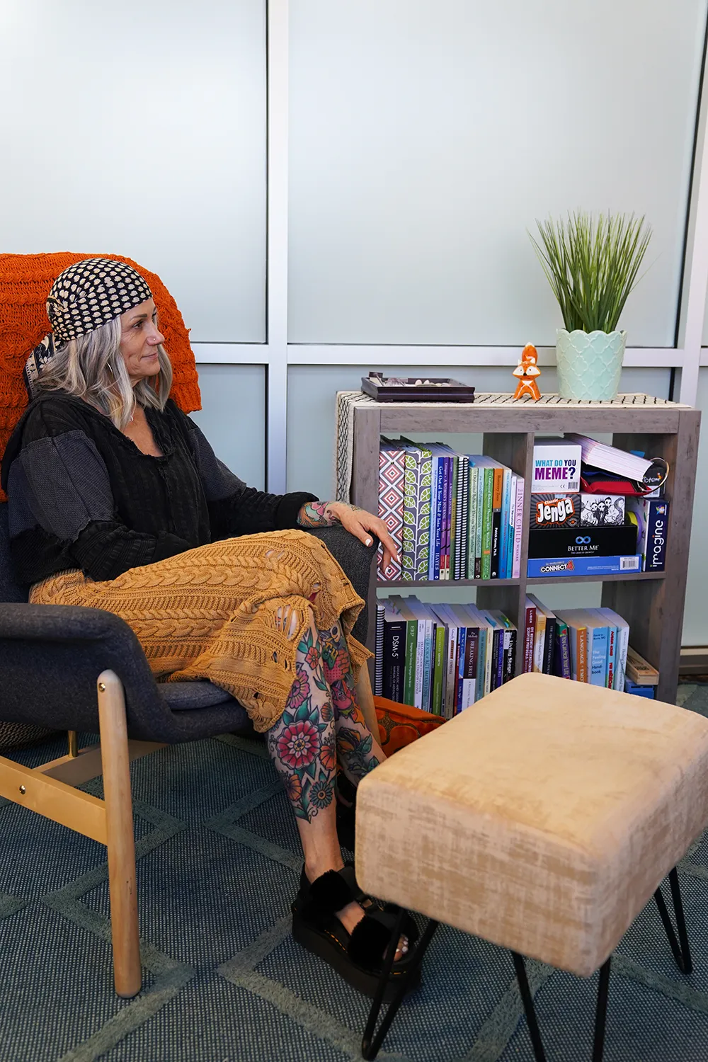 A woman seated in a wood-framed chair under a knitted orange throw, beside a bookshelf of games and clinical texts and an upholstered ottoman.