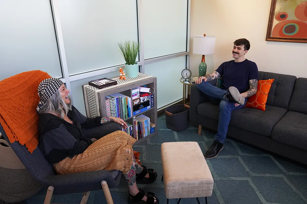 Two people in conversation across a grey sofa and a wood-framed chair.