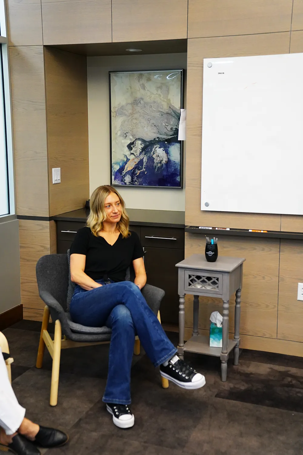 A staff member in an office chair beside a whiteboard, with a blue abstract canvas and a side table.