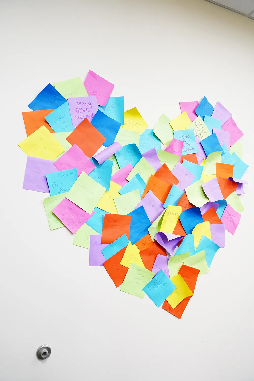 A heart made from dozens of coloured sticky notes on a white wall, each one handwritten.