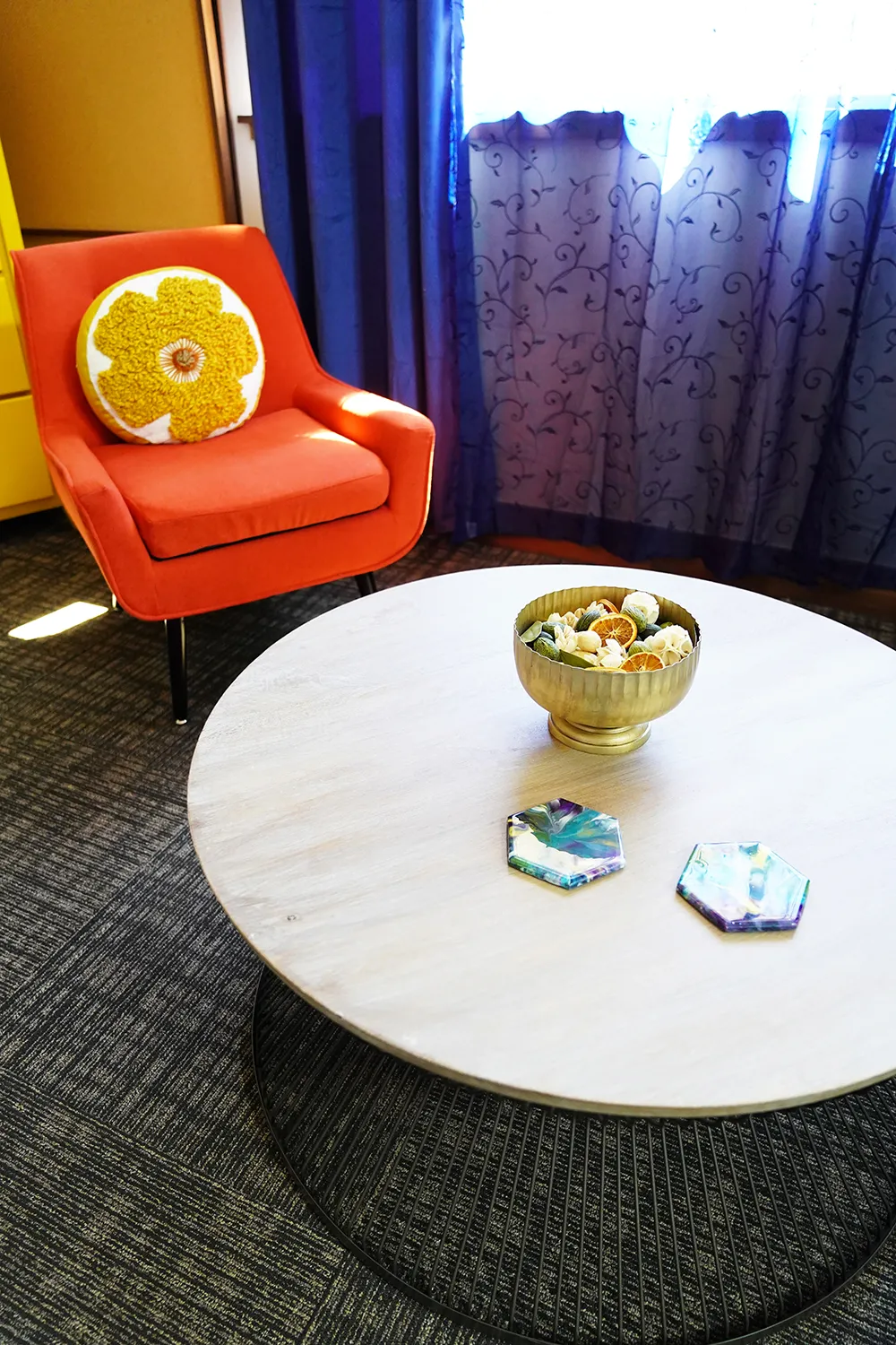 A corner with an orange armchair and citrus cushion, a round marble table holding a gold bowl, and a purple curtain.