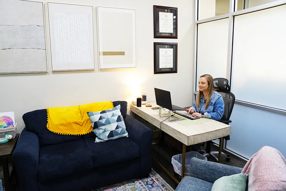 An office with a navy sofa, yellow and patterned cushions, framed art, and a staff member at a desk.