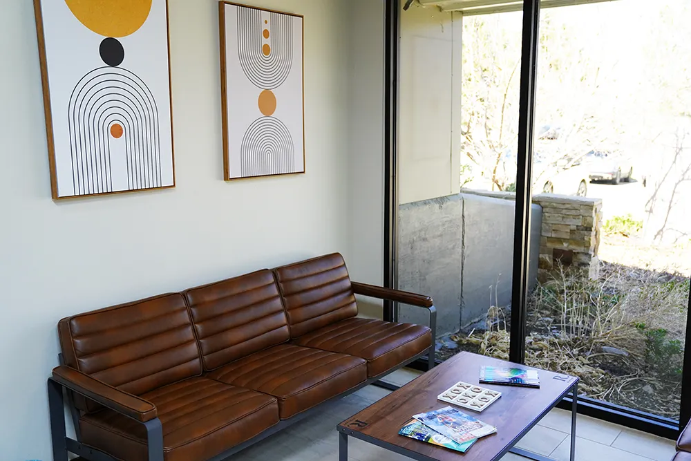 A brown leather bench beneath framed arch prints, beside floor-to-ceiling windows onto rock and greenery.