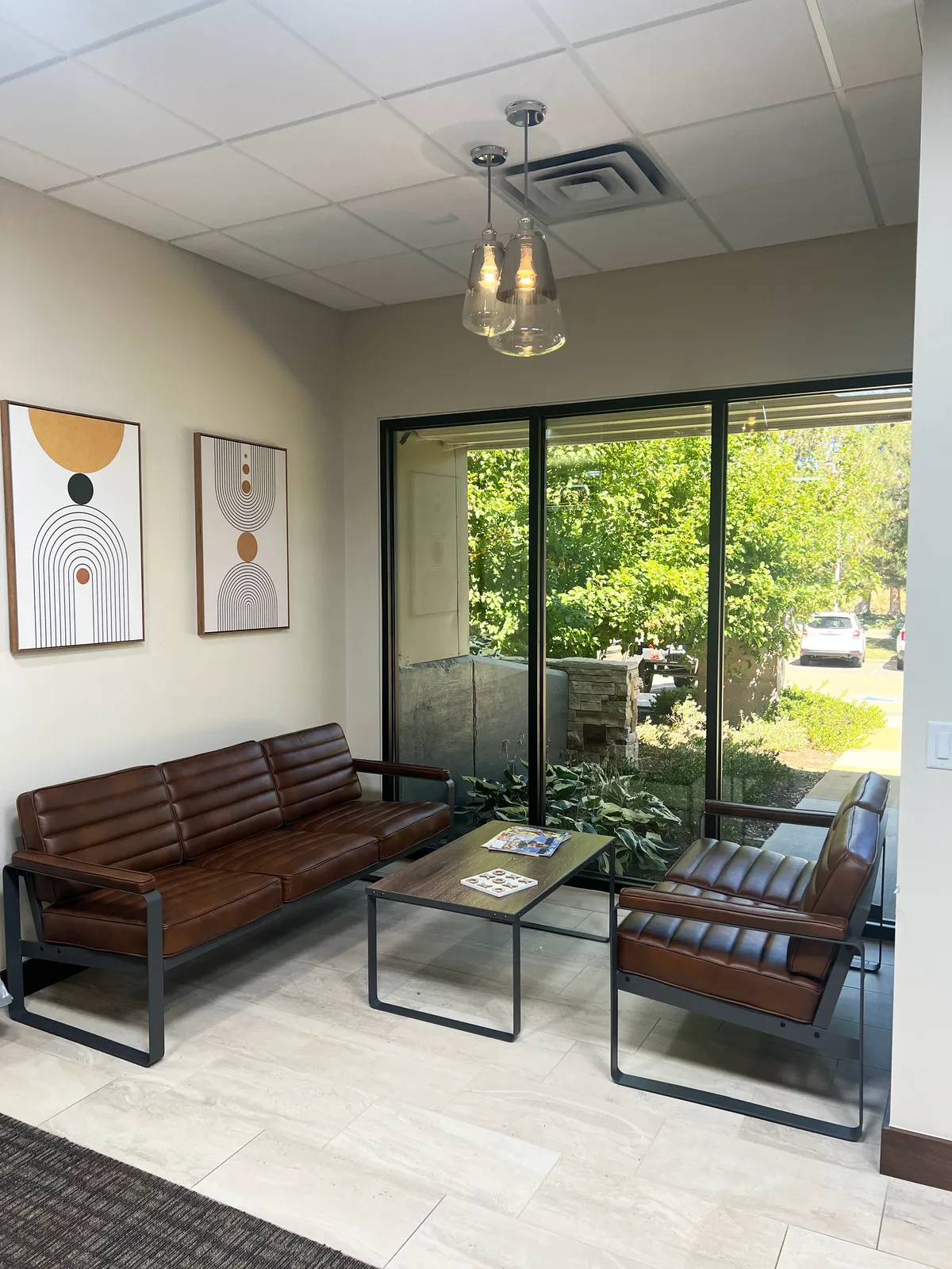 A waiting area with a brown leather sofa and armchair beside floor-to-ceiling windows looking out to green trees.
