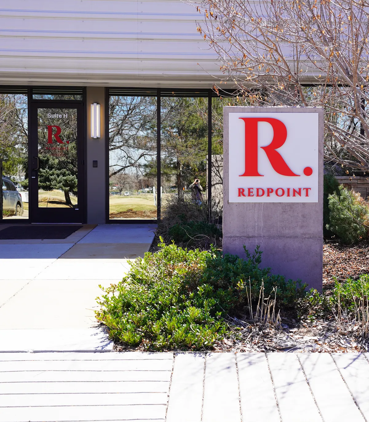 The entrance to The Redpoint Center's Longmont office: a glass door lettered Suite H beside a concrete monument sign carrying the red R. Redpoint mark, winter branches overhead.