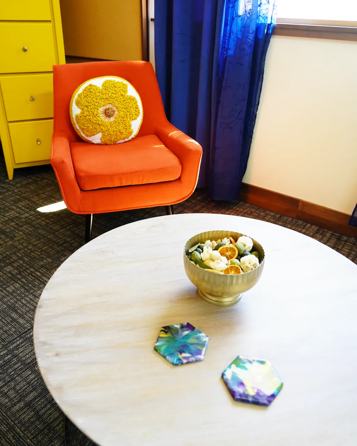 A burnt-orange armchair with a yellow flower cushion beside a round table holding a brass bowl of potpourri and two marbled coasters.