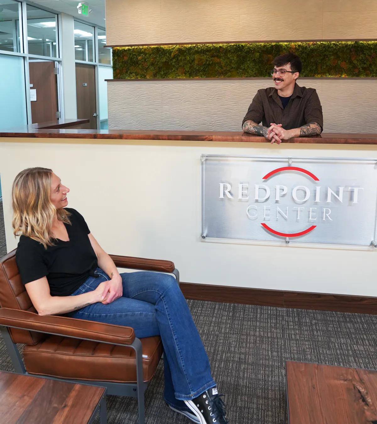 A Redpoint Center team member at the front desk welcoming a client seated in the Fort Collins reception area, the Redpoint Center sign mounted on the desk panel between them.