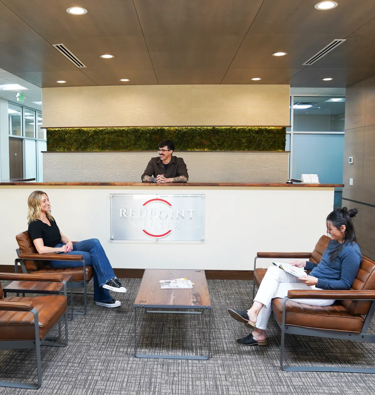 A Redpoint receptionist at the front desk smiling toward a woman seated in a leather chair, another client reading nearby, a moss wall behind them.