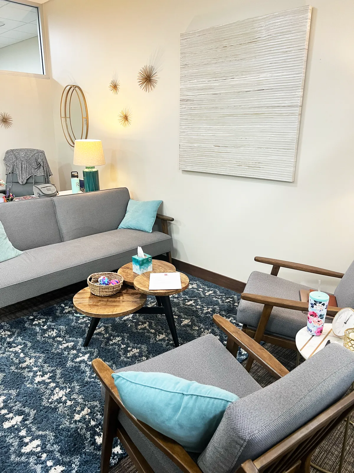An empty therapy room with a grey sofa and teal cushions, two wooden armchairs, and round wood tables holding a tissue box and a basket of fidget toys.