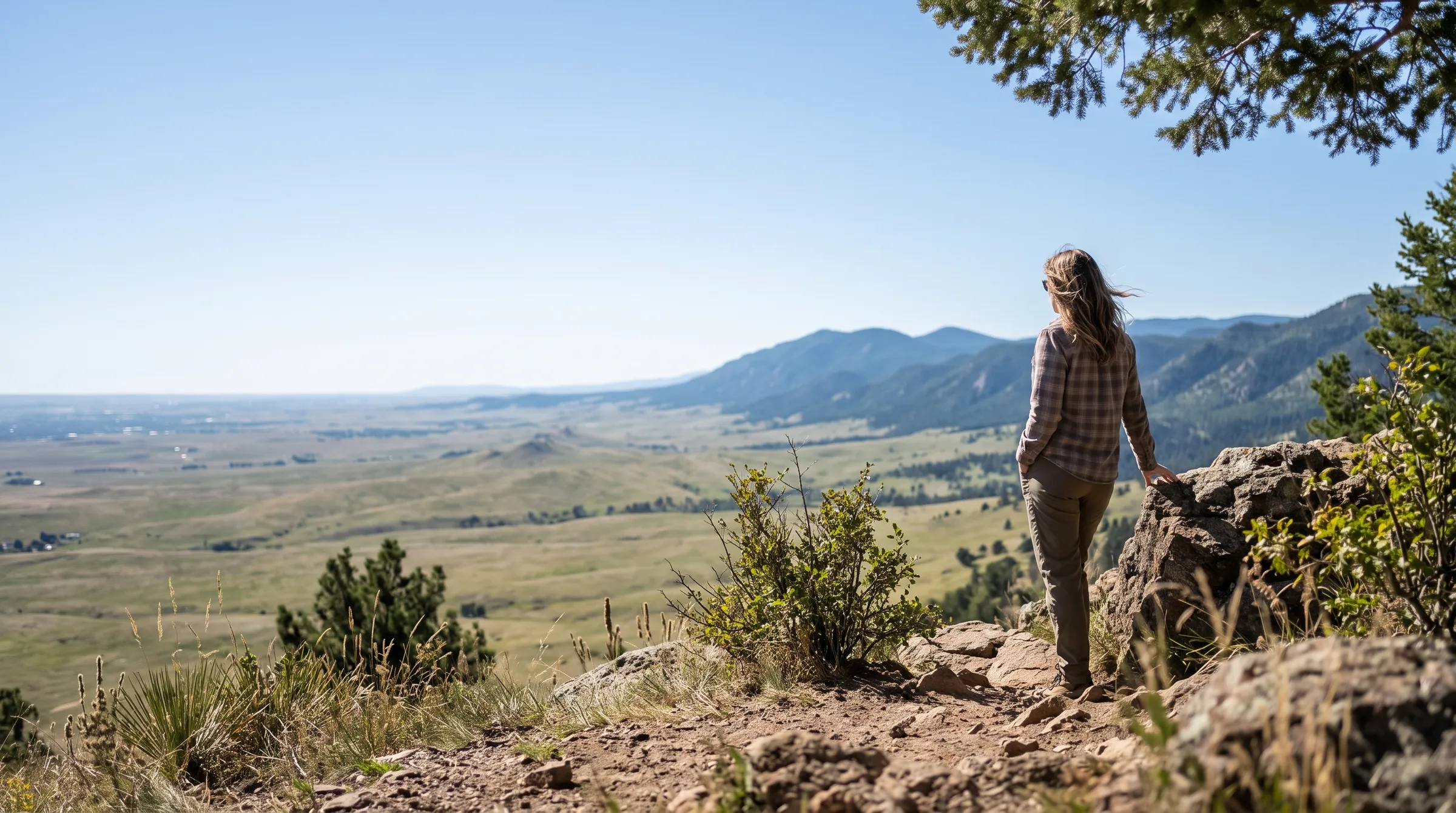 A person at a Front Range foothills overlook looking out over open grassland and soft blue mountains under a clear Colorado sky.