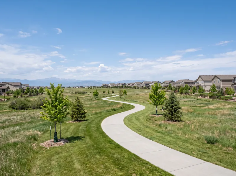 A concrete path winding through open prairie grass in a Thornton park, new suburban rooflines and the distant Front Range under a wide plains sky.