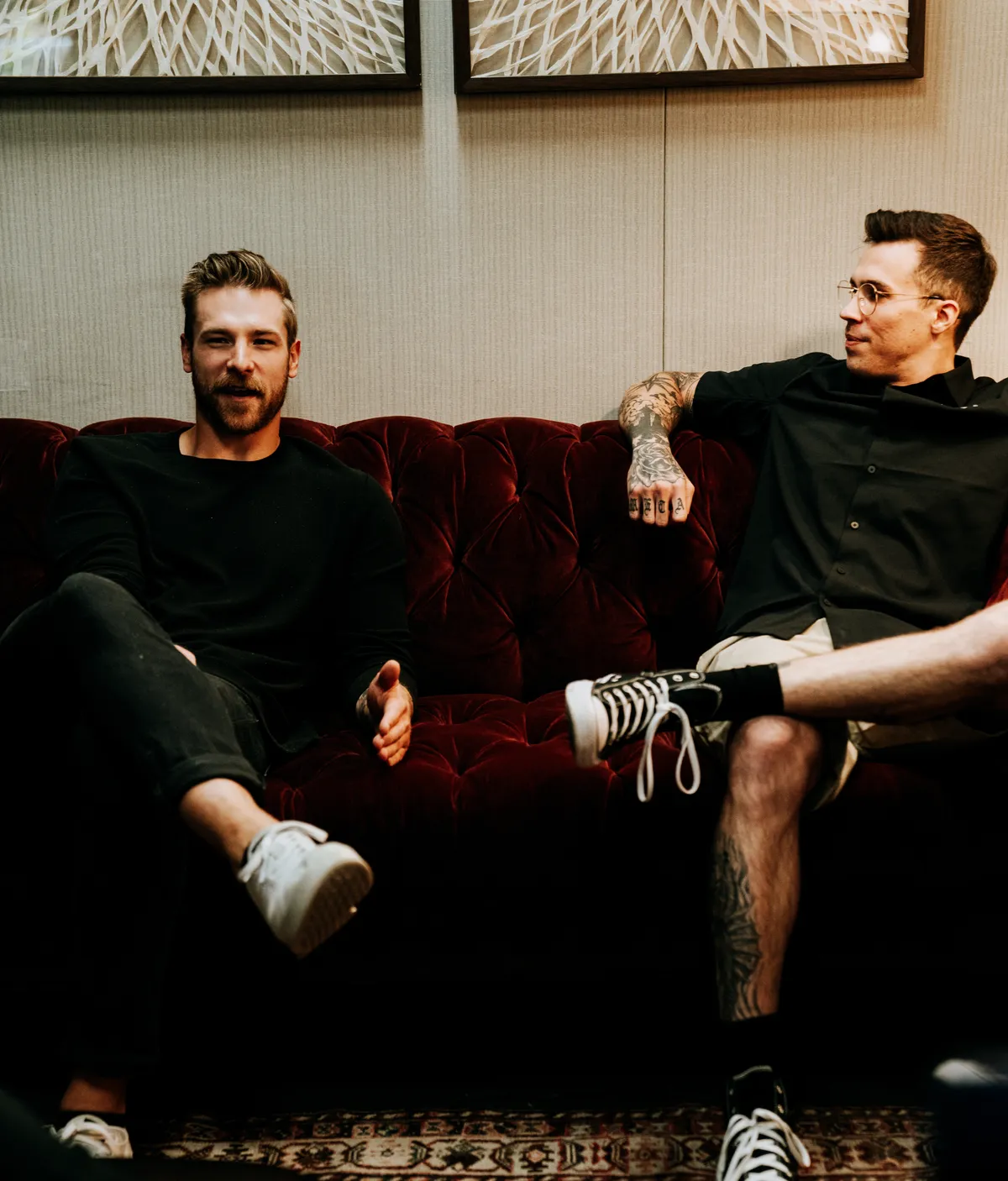 Two men on a deep red velvet couch in a book-lined lounge, mid-conversation.