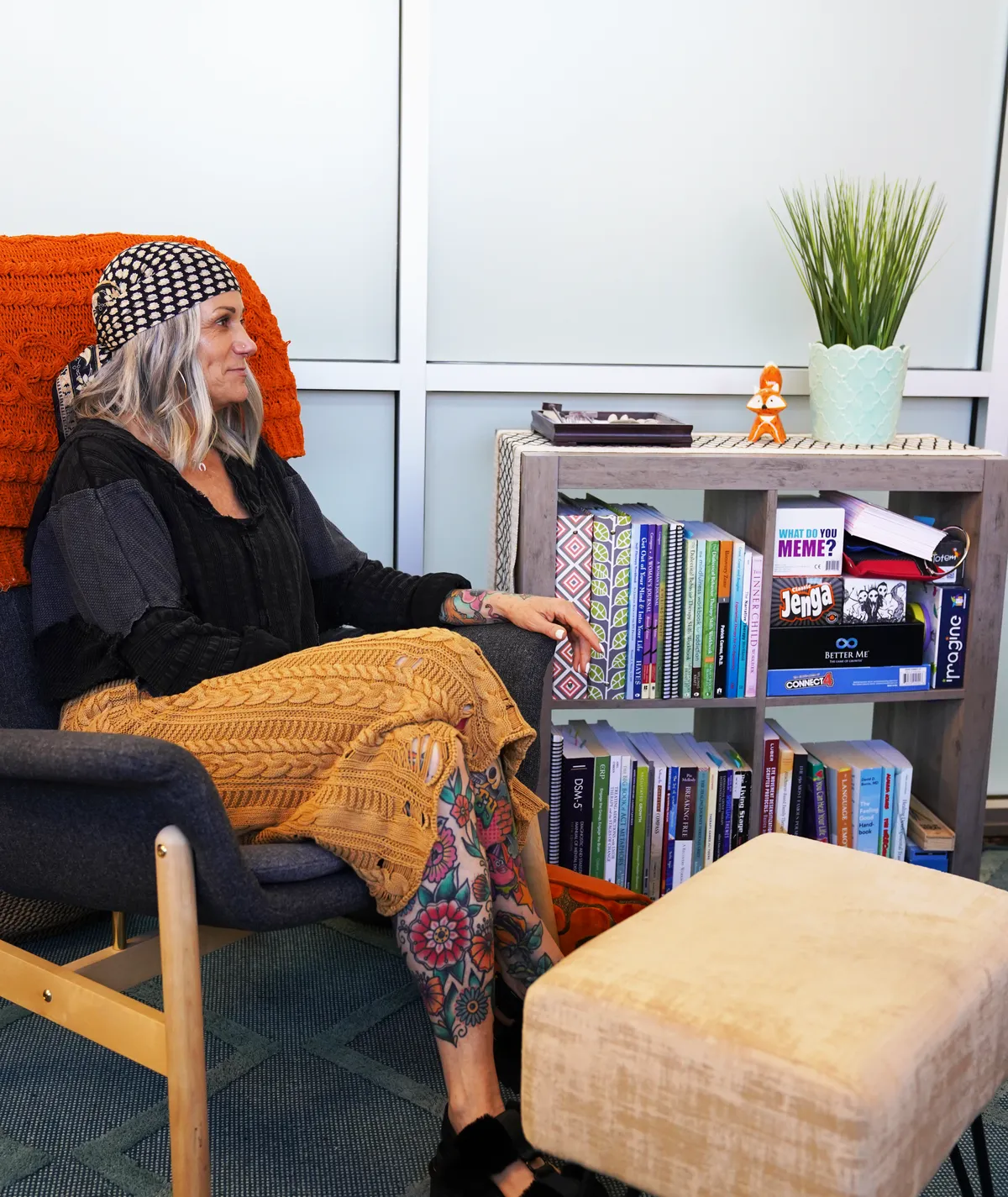 A woman in a patterned headscarf seated in a grey armchair beside a shelf of recovery workbooks and board games.