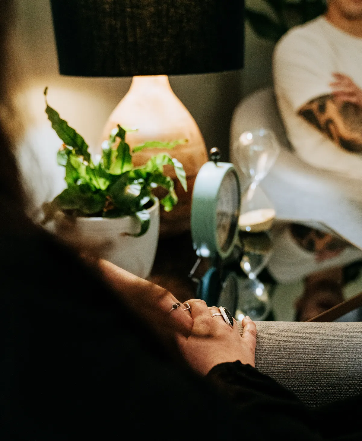 Clasped hands resting on a sofa arm beside a potted plant, a glowing ceramic diffuser, a small clock and an hourglass, a person soft-focus behind.