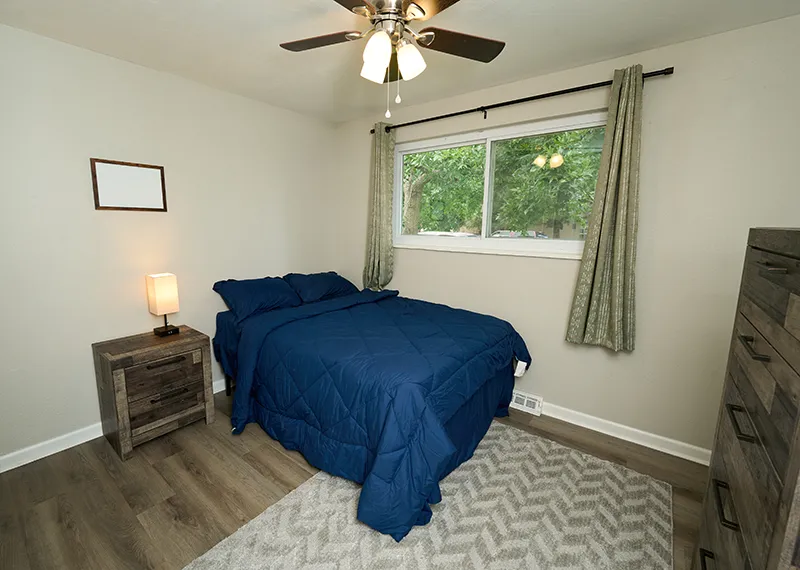 A bedroom with a bed in a blue comforter, a wood nightstand and lamp, framed art, a ceiling fan, and a window onto trees.