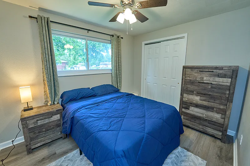 A bedroom with a bed in a blue comforter, a nightstand and lamp at left, white sliding closet doors, and a tall reclaimed-wood dresser at right.