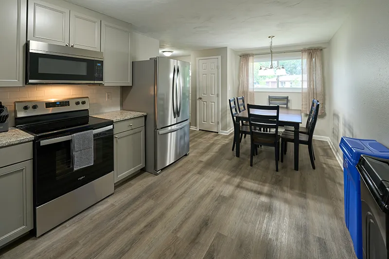 A kitchen with grey cabinets, a stainless range and refrigerator, opening through to a small dining table under a window.