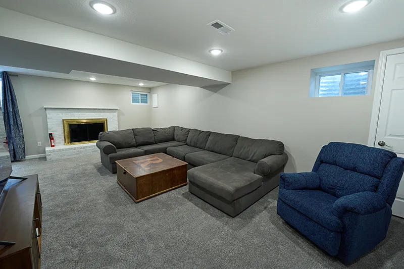 A garden-level living room with a white brick fireplace, a large grey sectional, a blue recliner, and a wooden chest used as a coffee table.