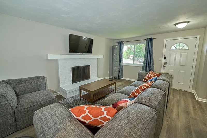 A front living room with grey sofas and orange patterned pillows, a white fireplace with a television above it, and a front door with a fanlight.