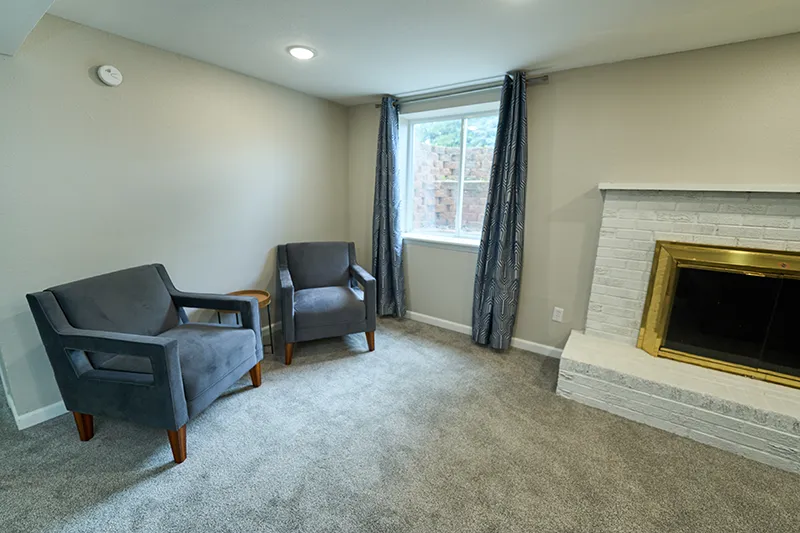 Two dark armchairs by a window beside a white brick fireplace with a brass surround.