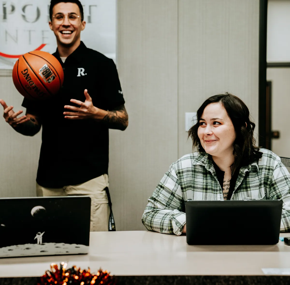 Two Redpoint staff laughing at a desk, one standing and tossing a basketball beneath the Redpoint banner.