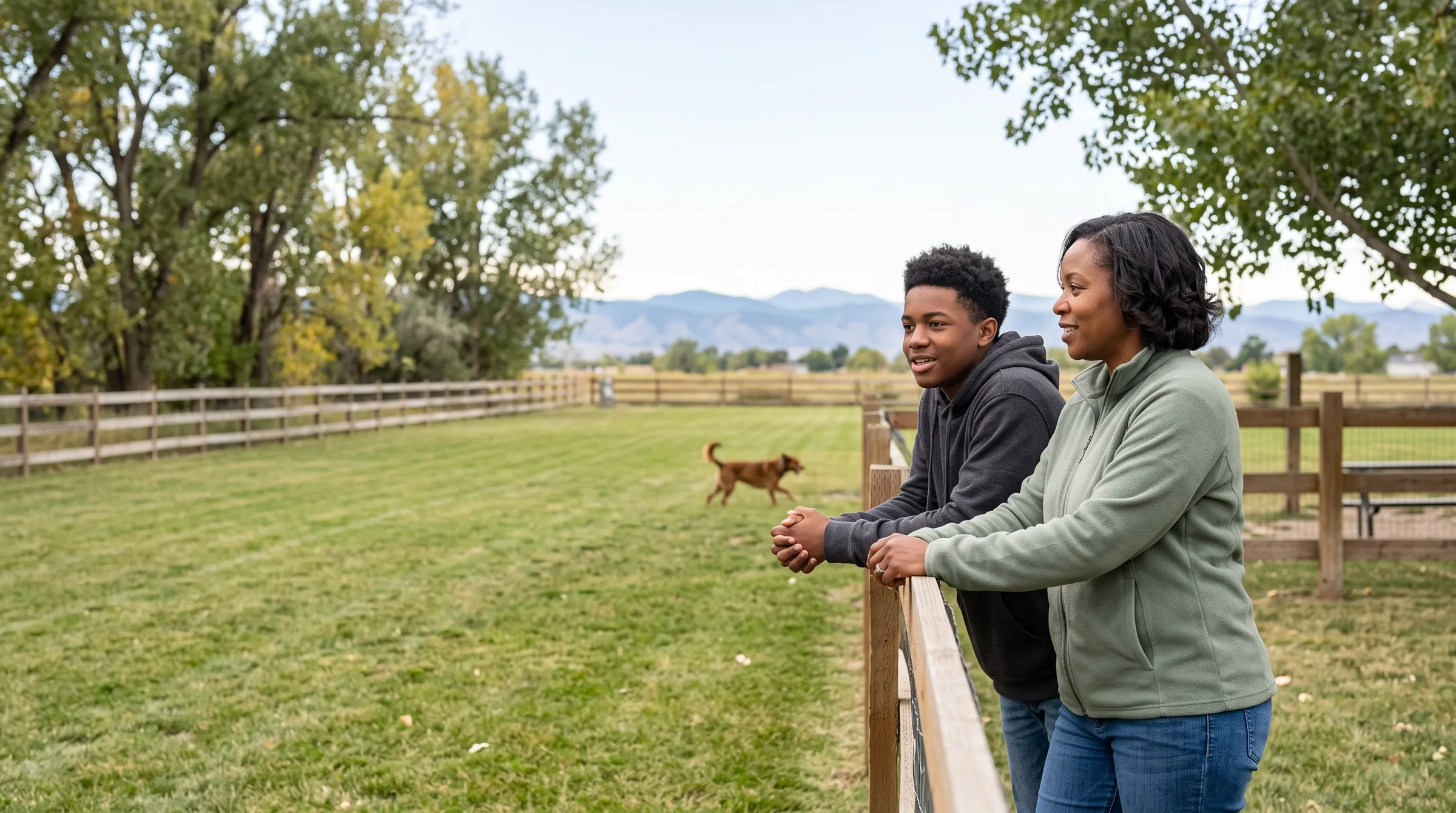 A mother and her teenage son leaning on the top rail of a dog park fence, looking out across the grass as their dog trots past in the middle distance.